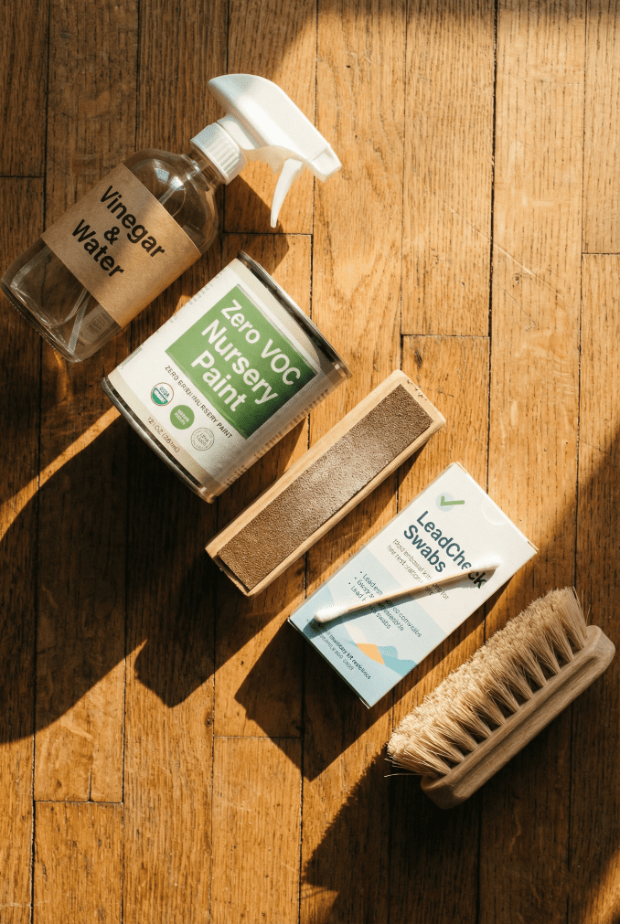 A flat lay of baby-safe cleaning supplies including vinegar, water, Zero VOC paint, and a lead test kit for restoring a used crib.