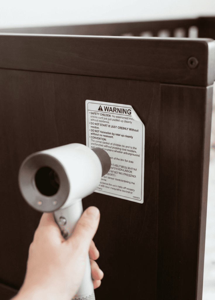 Delta Children Heartland 4-in-1 Crib Review: The Best Budget-Friendly & Safe Choice for 2026? 3 Close-up view of a hairdryer warming a safety sticker on the wood frame of a Delta Children Heartland crib to remove it easily.