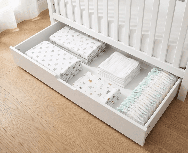 An open under-crib trundle drawer of the Davinci Colby crib, filled with folded crib sheets, burp cloths, and diapers, demonstrating storage space.