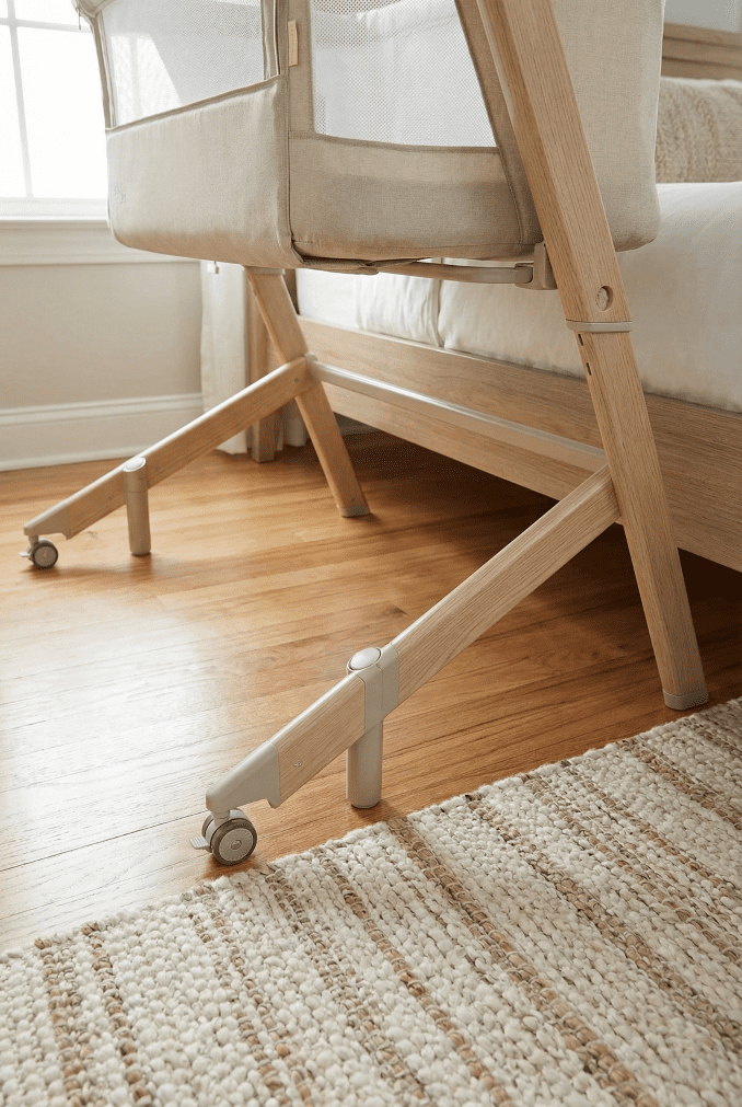 A low-angle view showing the stable, wood-finish metal legs of the Evolur bassinet sliding under an adult bed, highlighting its secure H-frame construction.