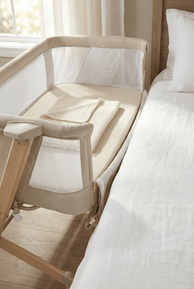 A close-up view of the Evolur Stellar bedside bassinet with its mesh side panel lowered next to an adult bed, demonstrating the easy access feature for parents.